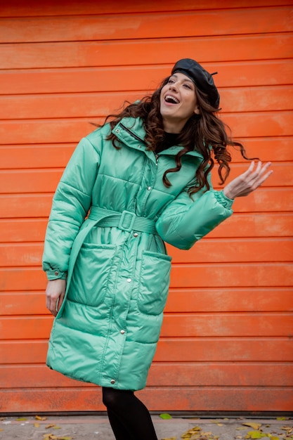 Free photo stylish woman posing in winter autumn fashion trend puffer coat and hat beret against orange wall in street