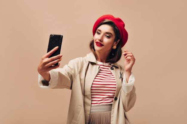 Stylish woman in red beret takes selfie on beige background. Beautiful lady with bright lipstick with earrings and in raincoat makes photo.