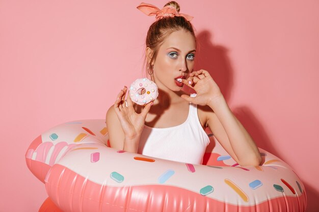 Stylish woman in round gold earrings and cool white swimsuit looking into camera holding donut and posing with swimming ring on pink backdrop