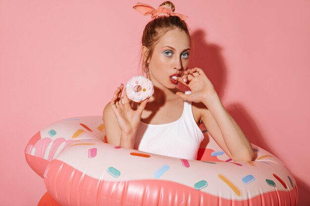 둥근 금 귀걸이와 멋진 흰색 수영복을 입은 세련된 여성이 도넛을 들고 분홍색 배경에서 수영 반지와 함께 포즈를 취하는 카메라를 바라보고 있습니다.