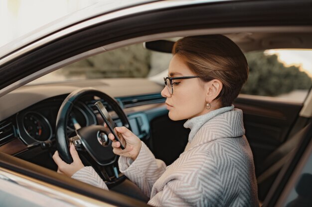 コートの冬のスタイルとスマートフォンを使用してメガネに身を包んだ車に座っているスタイリッシュな女性、忙しい運転のビジネスウーマン