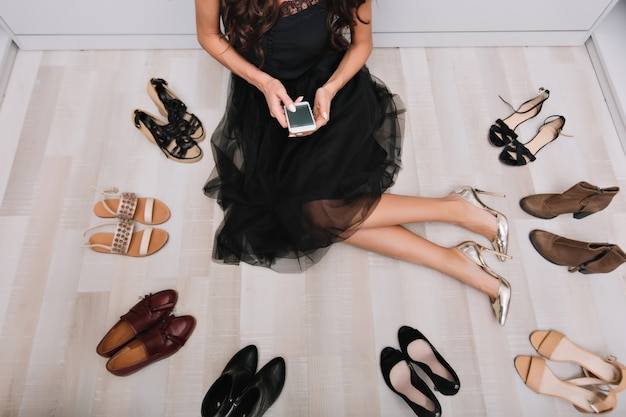 Stylish woman sitting on the floor in wardrobe with smartphone in hands, writing message, surrounded by lot of shoes. She wearing black fluffy skirt and silver luxury shoes.