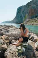 Free photo stylish woman sitting on rock holding palm leaves while taking photo with camera