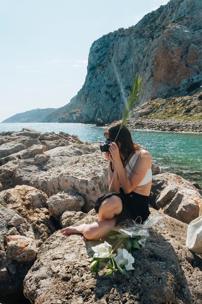 Стильная женщина, сидя на скале, держа пальмовые листья, принимая фото с камерой