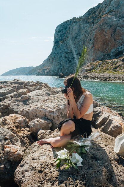 カメラで写真を撮っている間ヤシの葉を保持している岩の上に座っているスタイリッシュな女性