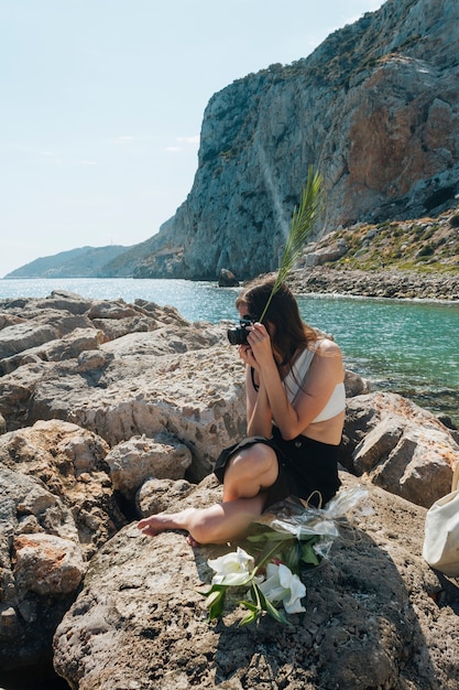 カメラで写真を撮っている間ヤシの葉を保持している岩の上に座っているスタイリッシュな女性