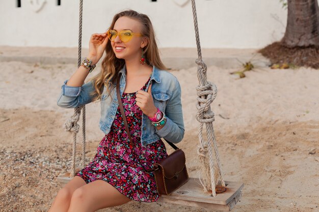 Стильная женщина в летнем платье отдыхает на пляже в красочных желтых солнцезащитных очках, улыбаясь счастливой