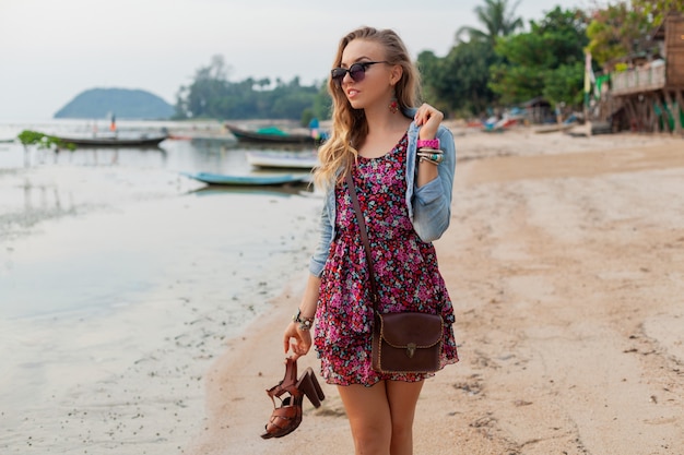 Стильная женщина в летнем платье на каникулах гуляет по пляжу с обувью в руке
