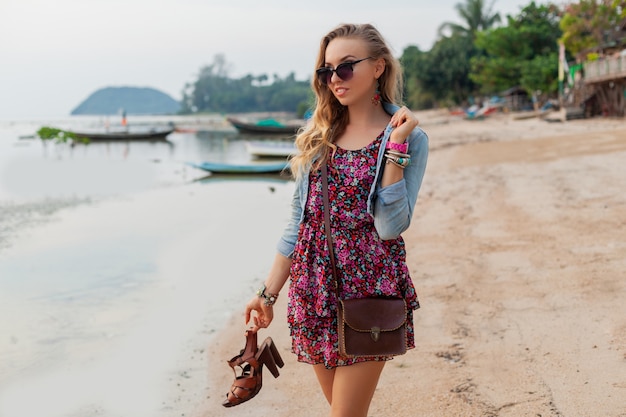 Стильная женщина в летнем платье на каникулах гуляет по пляжу с обувью в руке