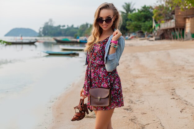 手に靴を持ってビーチを歩く夏のドレス休暇のスタイリッシュな女性