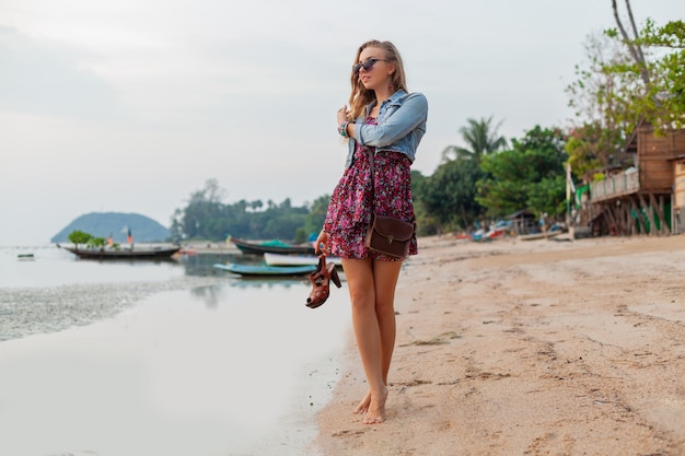 Stylish woman in summer dress vacation walking on beach with shoes in hand