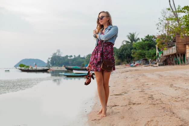 Stylish woman in summer dress vacation walking on beach with shoes in hand