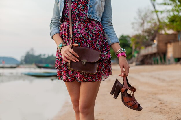 Stylish woman in summer dress vacation walking on beach with shoes in hand