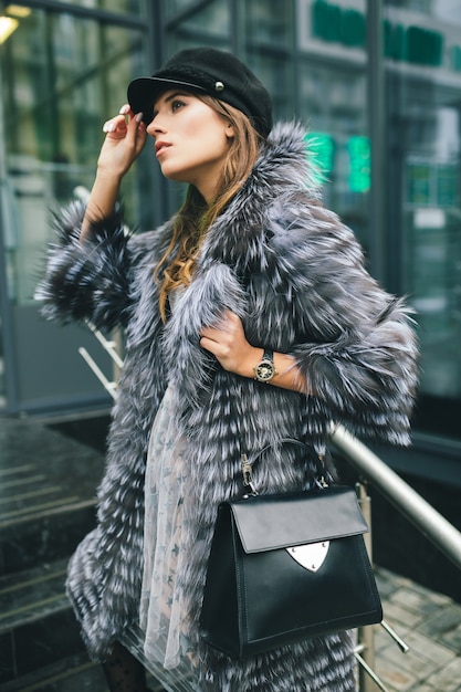 Stylish woman walking in city in warm fur coat, winter season, cold weather, wearing black cap, holding leather bag, street fashion trend, urban look