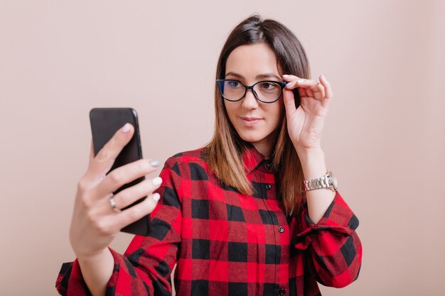 Stylish woman wears glasses making selfie on smartphone with happy emotions os isolated wall. Young woman using gadget with true emotions on isolated wall, place for text