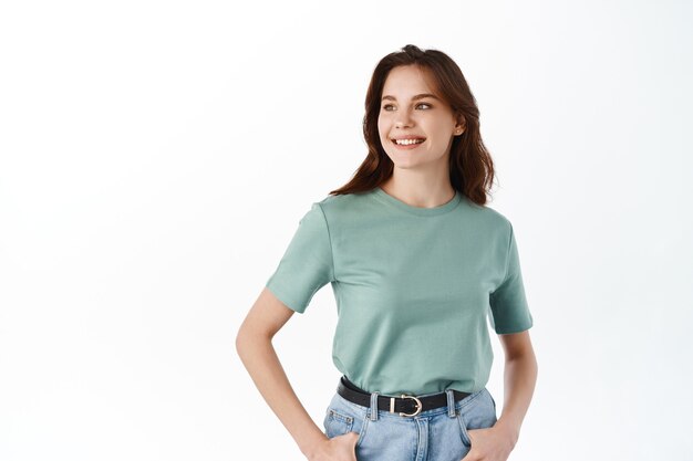 Stylish woman with confident and determined smile, holding hands in pockets of jeans, look aside and smile pleased, standing against white wall