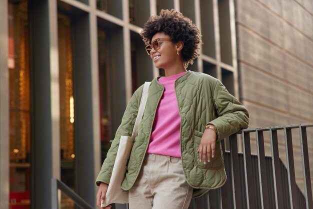 곱슬머리를 한 세련된 여성은 트렌디한 선글라스 재킷을 입고 바지는 패브릭 가방을 들고 도시 건물 근처에서 야외에서 즐거운 표정 포즈를 취하며 도시 사람들과 스타일 개념에서 자유 시간을 보낸다