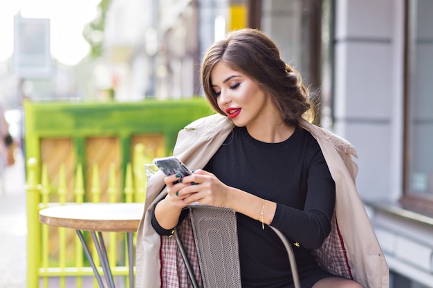 Stylish woman with red lips wearing beige coat scrolling smartphone while sitting in outdoor cafeteria
