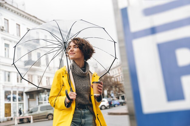 テイクアウトコーヒーを手に保持している大きな透明な傘の下で都市部を歩いて黄色のレインコートでスタイリッシュな女性