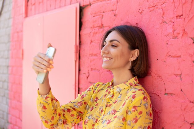ピンクのレンガの壁に黄色の夏のドレスを着たスタイリッシュな女性幸せなポジティブ携帯電話で自分撮り