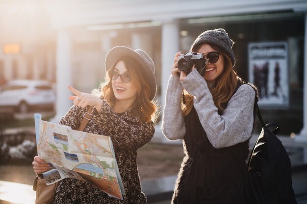 Стильные женщины проводят время на свежем воздухе в холодный день, исследуя новые места с камерой. Великолепная женщина-фотограф гуляет по городу с сестрой, которая показывает пальцем и улыбается, держа карту.