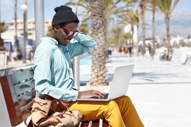 Stylish young African American male freelancer wearing hat and shades using laptop for remote work, using free city wireless internet connection, sitting alone on bench on promenade by the sea