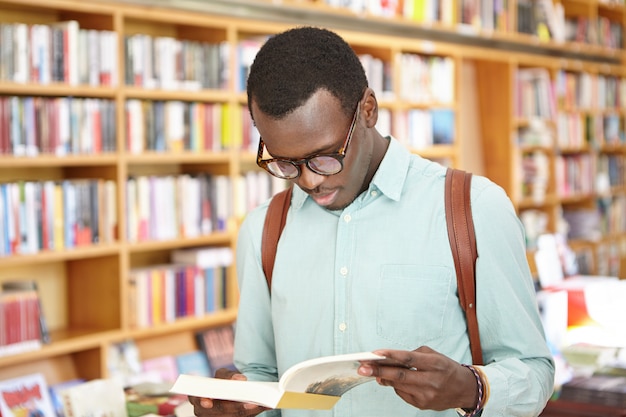 Stylish young African American male in shirt and eyewear looking through book in bookstore standing. Black male tourist exploring local bookshops while traveling abroad