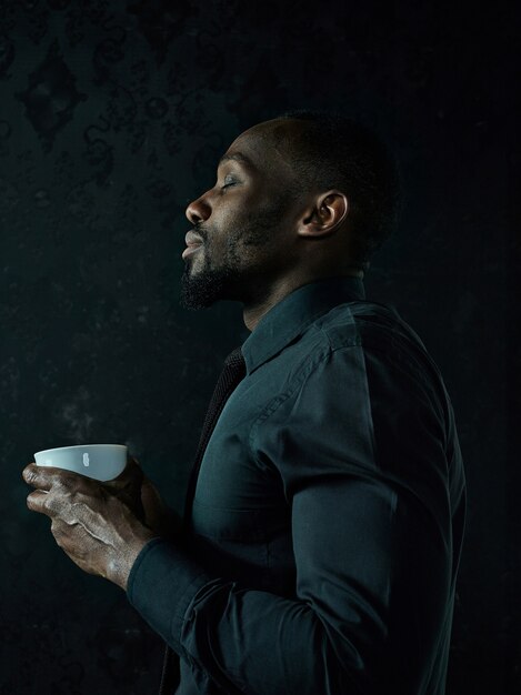 暗いスタジオの背景にポーズをとって白いコーヒーカップとスタイリッシュな若いアフリカの黒人男性。
