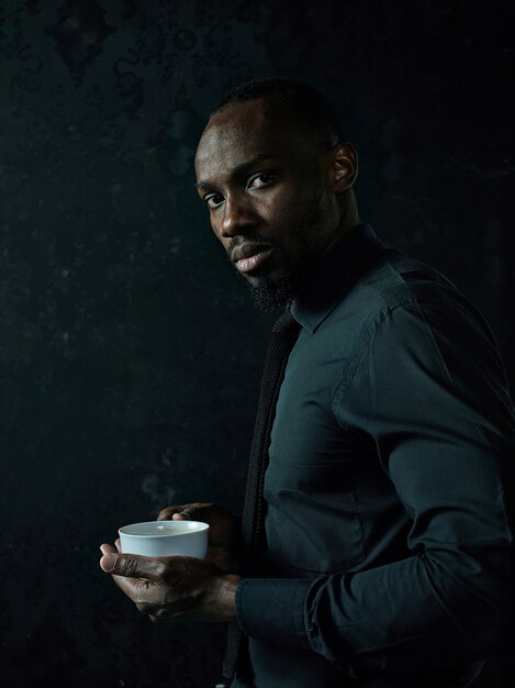 Стильный молодой африканский темнокожий мужчина с белой чашкой кофе позирует на темной студии.