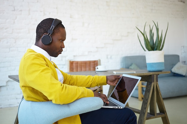 Stylish young afro american man sitting in chair at home, multitasking, using laptop and headphones, having serious facial expression. people, technology, communication and modern lifestyle