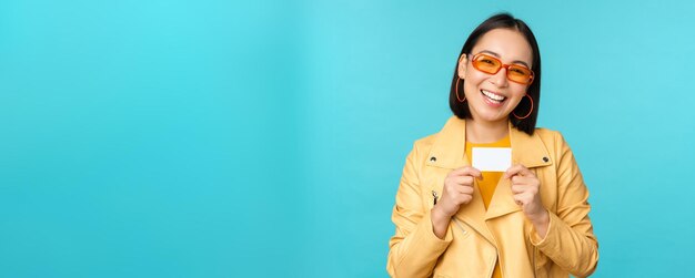 Стильная молодая азиатка в солнечных очках показывает кредитную карту и улыбается, рекомендуя банковские бесконтактные платежи или скидки в магазине, стоящем на синем фоне