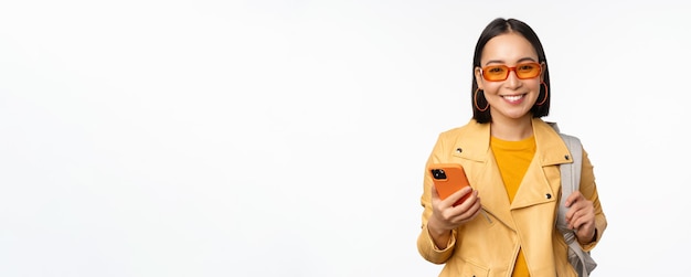 Stylish young asian woman tourist traveller with backpack and smartphone smiling at camera posing ag