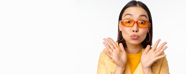 Stylish young asian woman in trendy sunglasses pucker lips silly looking surprised impressed reaction standing over white background