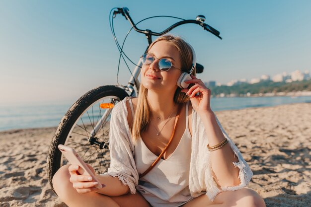 音楽を聴いてヘッドフォンで自転車とビーチに座っているスタイリッシュな若い魅力的な金髪の笑顔の女性