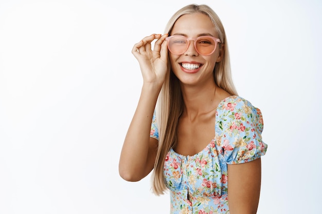 Stylish young blond woman in sunglasses smiling and looking positive at camera wearing dress standing over white background