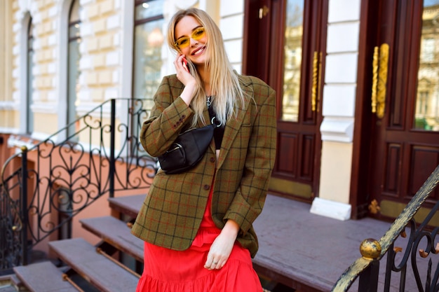 Stylish young blonde woman posing near luxury shop and speaking by her smartphone, fashionable modern outfit, oversized jacket and bum bag.