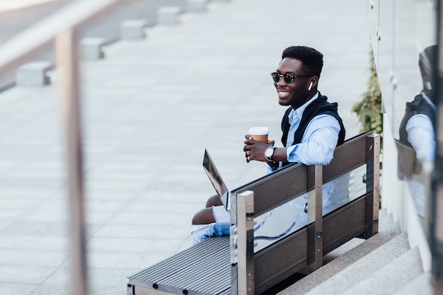 Stylish young businessman siting on a bench with his laptop on a sunny street next to a park. With cup of coffee. Lifestyle.