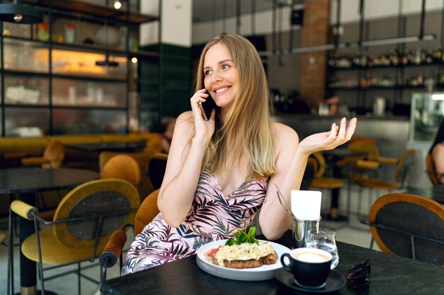 スタイリッシュな若い実業家がブランチカフェで屋内でポーズをとり、彼女のスマートフォン、エレガントな衣装をすすりながら。