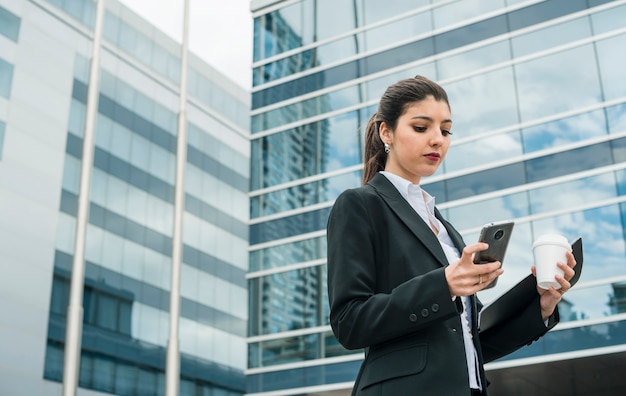 Stylish young businesswoman standing in front of corporate building texting message on mobile phone