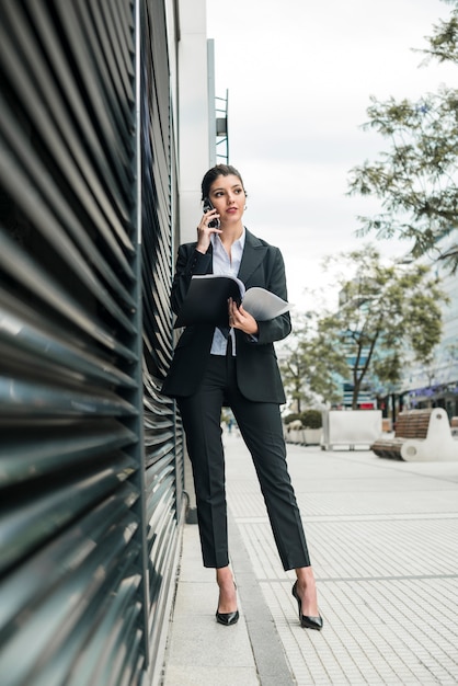 Free photo stylish young businesswoman talking on mobile phone holding folder in hand