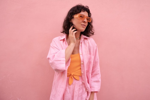 Stylish young caucasian brunette woman in glasses summer clothes looks away spends time on pink background