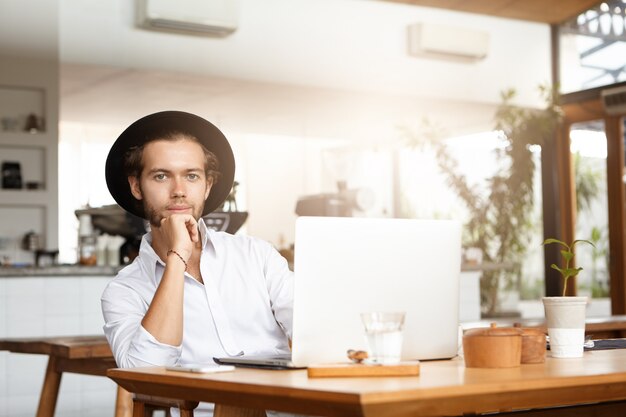 졸업장 프로젝트에서 작업하는 동안 나머지를 갖는 검은 모자에 세련된 젊은 백인 학생, 열린 노트북 컴퓨터 앞에 카페 테이블에 앉아 그의 팔꿈치에 기대어 미소로보고