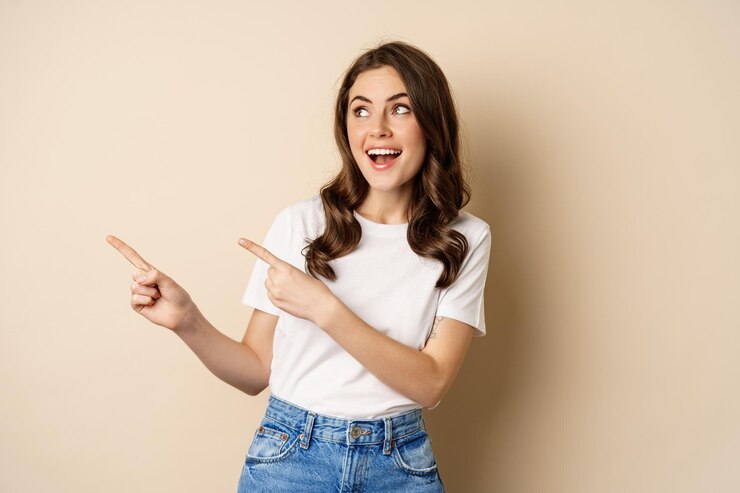 Free Photo stylish young caucasian woman smiling, pointing fingers left, showing advertisement, promo offer, standing against beige background.