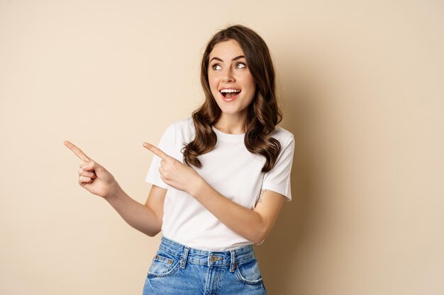 Stylish young caucasian woman smiling, pointing fingers left, showing advertisement, promo offer, standing against beige background.