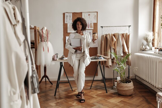 Stylish young curly dark-skinned woman in white pants, jacket and blouse leans on desk in fashion designer office, holds coffee cup and laptop