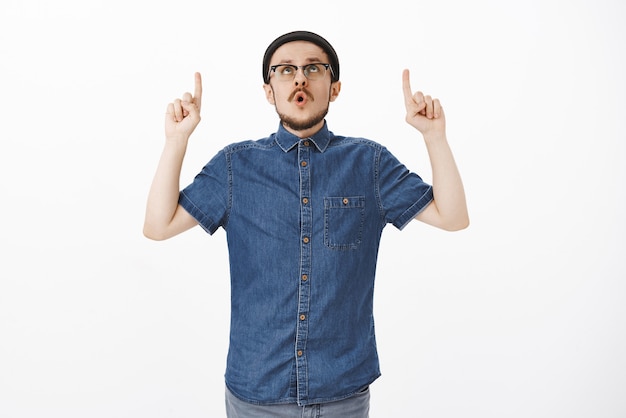 Stylish young european male with beard and moustache in hat and glasses folding lips saying wow pointing and staring up excited over white wall
