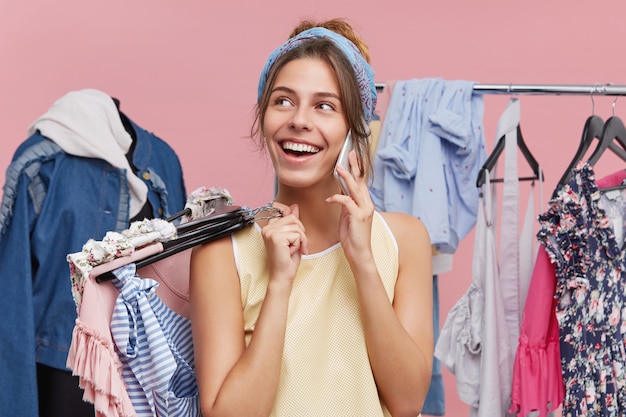 Elegante giovane femmina shopaholic che parla al cellulare con la sua amica, vantandosi dei suoi acquisti mentre fa shopping nel centro commerciale della città, in piedi a rack pieno di colorati capi alla moda