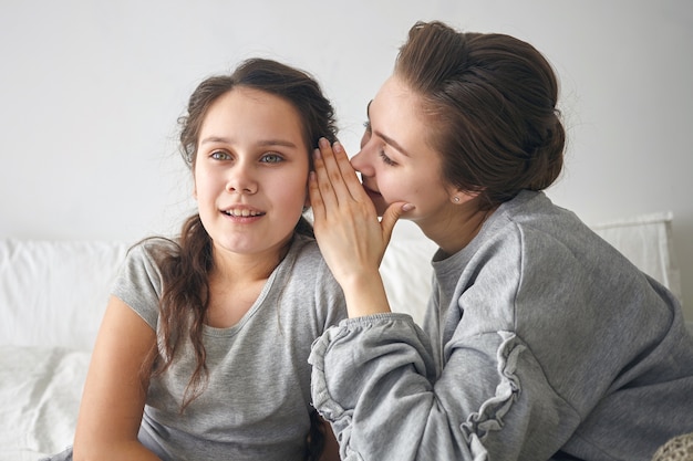 Free photo stylish young female whispering something into ear of her fascinated little sister, sharing secret.