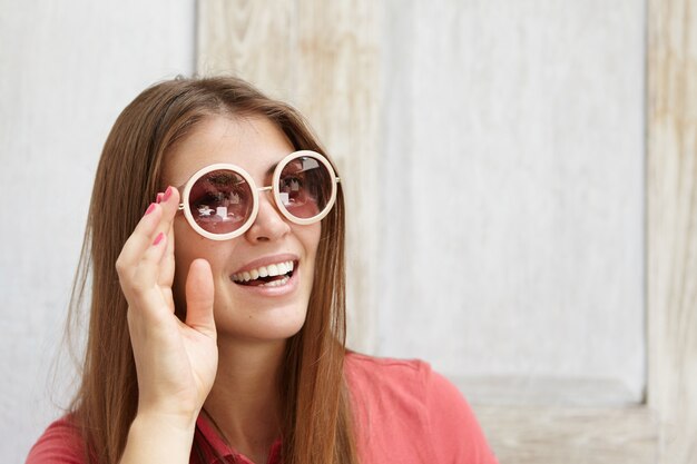 Stylish young female with pink nails adjusting her round sunglasses while relaxing indoors on sunny day. Pretty student girl with happy smile spending morning at home before going out to college