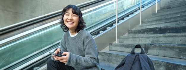 Stylish young girl student sits on stairs with smartphone and backpack laughs and smiles texts